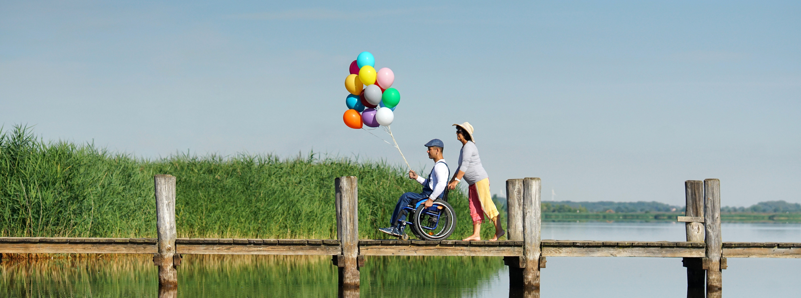 To smilende mennesker på vej hen ad en badebro. Solen skinner og der er vand under broen og siv i
baggrunden. Manden sidder i kørestol og har 14 balloner i forskellige farver i snore svævende over sit
hoved. Kvinden skubber kørestolen.